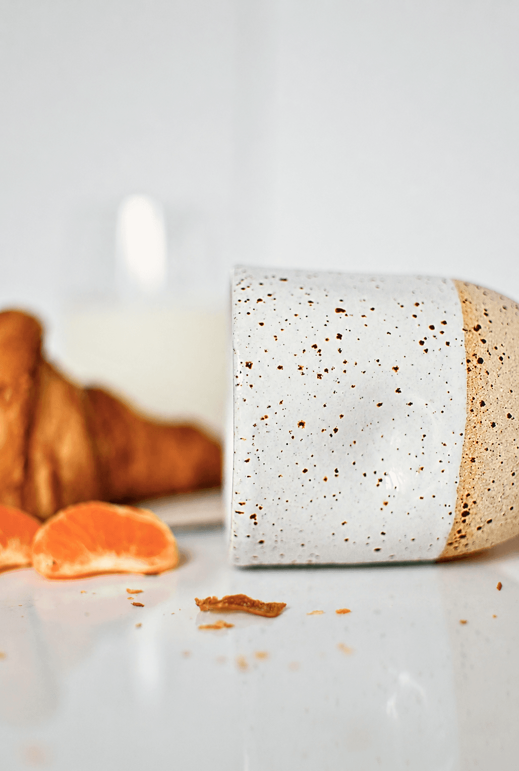 Side view of a ceramic coffee cup, wheel-thrown in speckled stoneware and glazed in glossy white. A fresh croissant, a glass of milk and mandarine wedges surround the cup, showcasing a typical morning breakfast.