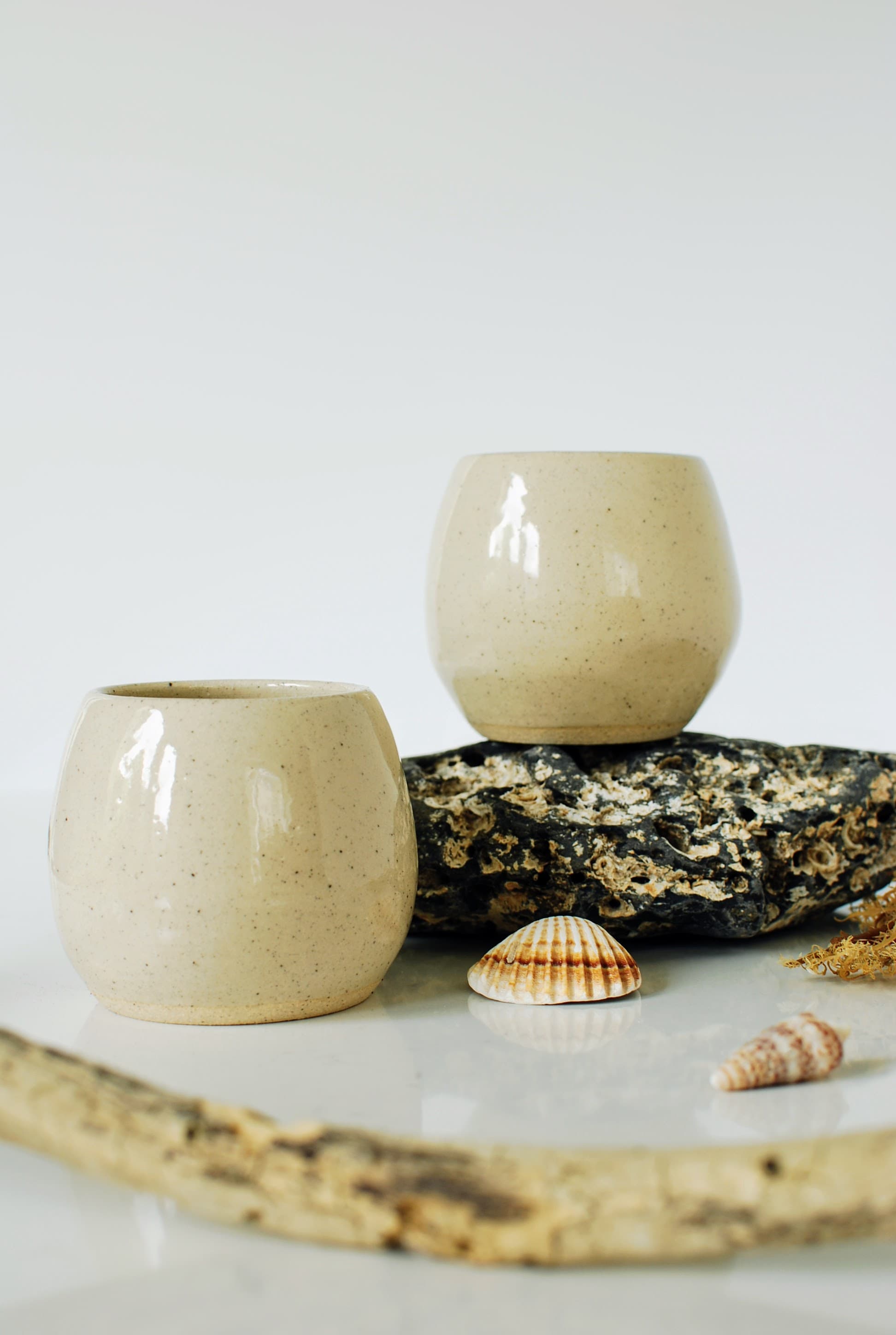 Handmade stoneware coffee cups in warm sand glaze, styled on coastal rock with seashells β Sable collection, wheel-thrown pottery made in Dublin, Ireland