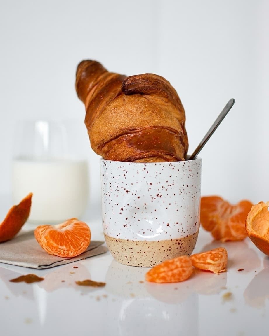 Side view of a ceramic coffee cup, wheel-thrown in speckled stoneware and glazed in glossy white. A fresh croissant, a glass of milk and mandarine wedges surround the cup, showcasing a typical morning breakfast.