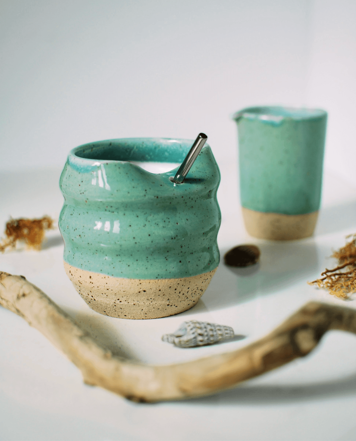 Handmade stoneware tumbler with organic rippled form and aqua turquoise glaze, styled with stainless steel straw and dried botanicals β Maguelone collection, made in Dublin, Ireland